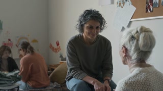 Medium full shot of two female natural disaster survivors chatting in refugee shelter, mature Caucasian woman consoling senior lady with grey hair, young boy playing on bed in background