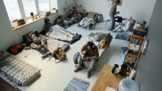 Wide high-angle surveillance camera shot of people displaced by natural disaster, flood or fire resting in emergency shelter – sleeping, pouring tea, listening to music, mom reading book to son