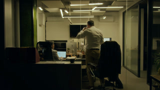 Full rear shot of middle-aged Caucasian male manager finishing work on report on computer late at night, getting up to go home, taking jacket, saying goodbye to Hispanic female colleague