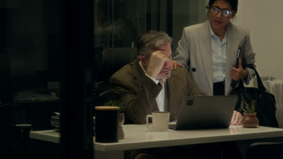 Medium shot of female colleague saying goodbye to exhausted workaholic male manager working overnight on project on laptop in dark office, patting on shoulder and urging to go home