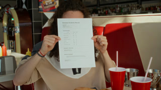 Medium portrait of nerdy gen Z Caucasian male student with curly long hair, eyeglasses covering face with exam grade sheet, lowering and pointing at successful score, while celebrating at burger diner