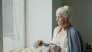 Medium shot of pensive elderly Caucasian female natural disaster survivor standing by window in refugee camp, drinking tea, reflecting on traumatizing experience and distressing memories, sighing