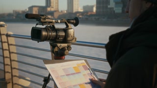 Medium midsection shot of anonymous male operator or videographer standing by camera on tripod on river bridge, studying broadcasting schedule on clipboard and circling next assignment