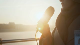 Medium closeup with lens flare of anonymous young female journalist in warm coat holding news channel microphone, delivering news report while standing on river bridge against setting sun