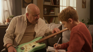 Medium shot of middle aged music teacher explaining electric guitar to young boy taking notes during music lesson at home scene lit by warm lighting Ideal for structured lesson promotion for kids