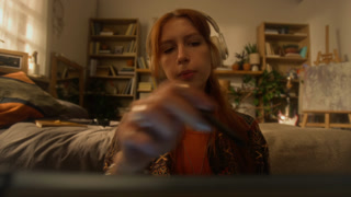 POV shot of red haired young woman wearing big headphones and listening to music while drawing on digital tablet screen working on creative projects at home in eclectic interior, copy space