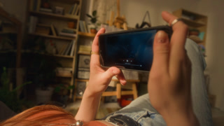 Over shoulder shot of young woman watching videos online and holding smartphone in hand while lying on bed at home relaxing, zoom in on smartphone screen with star constellations