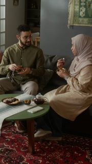 Vertical shot of young Muslim family enjoying tea time together in cozy modern home with woman wearing traditional clothing pouring tea for husband