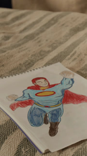 Vertical close-up of sketchbook with kids drawing of comics superhero in red cape and blue costume flying in air with clenched fists on bed in boy room, colored in pencil, creative hobby concept