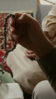 Vertical closeup shot of anonymous Muslim man holding beads in hand and praying at home with ornate rug on floor in background