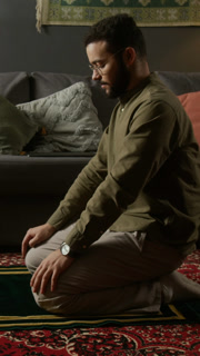 Vertical side view shot of adult Muslim man kneeling on prayer mat and praying with eyes closed while doing daily religious ritual in home living room