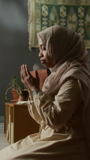 Vertical side view shot of Muslim young woman wearing hijab finishing daily prayer ritual at home turning head during Salah in calm home setting