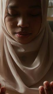 Vertical zoom in shot of smiling Muslim young woman wearing headscarf and praying at home soft light with shadows accentuating face and natural beauty