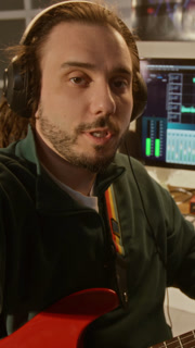 POV of Caucasian young adult man wearing headphones, holding electric guitar, speaking to camera in home studio. Video is ideal for online music tutorials, podcast or creative vlogs, vertical