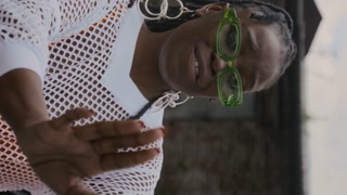 Vertical handheld shot of young African American lady in glasses and classy outfit rapping to camera while filming music video in shabby area