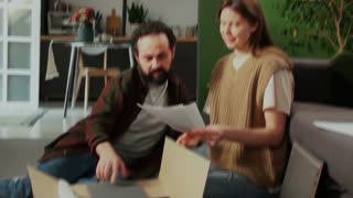 Medium handheld shot of affectionate, loving mature Caucasian couple unpacking DIY assembly drawer, discussing instructions, hugging and looking at camera, family life moments filmed on camcorder