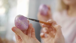 Close up of female hands using brush and decorating purple Easter egg with golden glitter