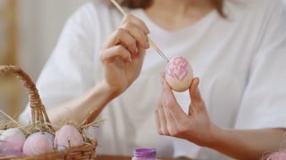 Handheld close up shot of hands of unrecognizable woman using brush and painting pattern with purple gouache on Easter egg