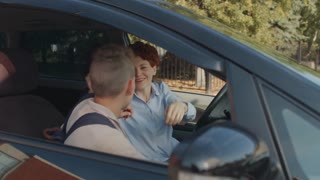 Handheld tracking of cheerful mother with short red hair sitting in car and hugging her teen son with backpack while dropping him off at school