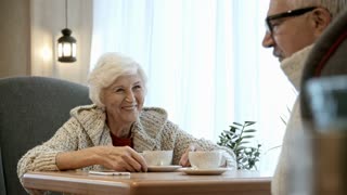 Joyous senior woman sitting at table in coffee shop, smiling and listening to elderly man telling a story
