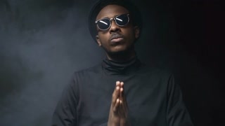 Low-key portrait shot of confident black man in hat and sunglasses standing in dark studio filled with smoke and bowing his head while holding his hands in prayer position