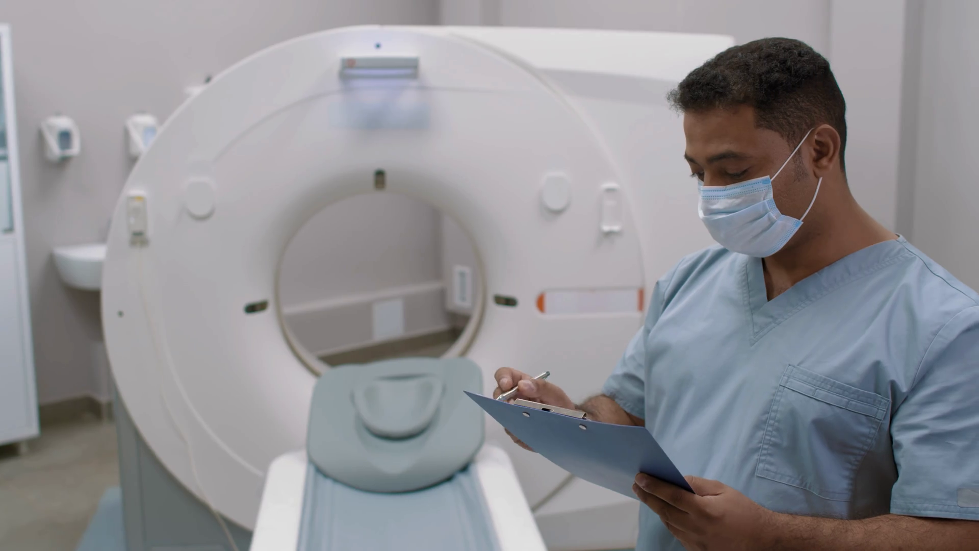 PAN of male technologist in face mask writing on clipboard in CT scan