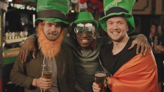 Slow-motion medium portrait of cheerful multi-ethnic men in funny green hats with clover posters celebrating St Patricks Day in pub, drinking beer and having fun together