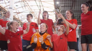 Team of happy female soccer players yelling in excitement, raising hands up and smiling while posing with cup in confetti shower after winning competition