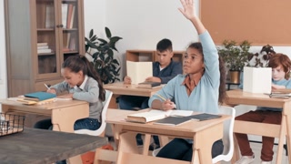 Tracking shot of smart African-American schoolgirl sitting at her desk and writing in notebook in classroom, then raising her hand and answering question