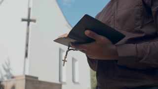 Medium close-up tilt down of African American pastor standing outdoors on summer day reading the Bible or prayer book