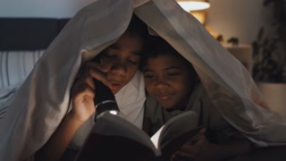 Medium close-up of cheerful Black girl and boy lying on stomachs under blanket on bed at night, reading book with flashlight, smiling and talking