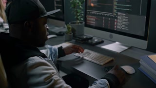 Medium close-up of young African American male mobile application developer in hoodie and baseball cap sitting at desk in office, writing computer code, then checking for errors on smartphone
