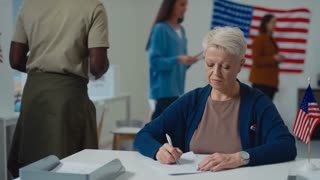 Medium portrait shot of smiling middle-aged Caucasian female municipal clerk working with ballot papers at electoral precinct during state referendum, with USA flag on table