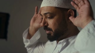 Side closeup of Biracial religious man in muslim cap performing takbirs and raising hands then bowing to Allah during namaz at home