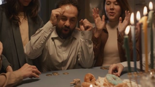 Medium shot of middle-aged Jewish man with beard, in kippah, playing fun traditional game with family at Hanukkah dinner, spinning dreidel, cheering, laughing and betting gelt coins