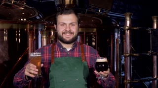 Happy male brewer in apron smiling joyfully holding two glasses of craft beer