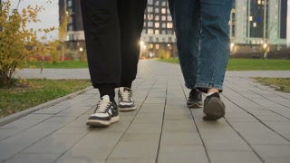 Leg view of two people walking side by side outdoors on paved path, one in black joggers and sneakers, other in jeans and black canvas shoes