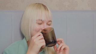 Blonde woman with bangs wearing green shirt smiles warmly while sipping from brown glass cup, she appears joyful and relaxed in soft indoor