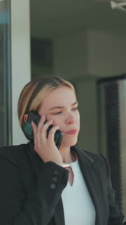 Frustrated woman during phone call lowers phone briefly, continues conversation after work while exiting office building, dressed in formal