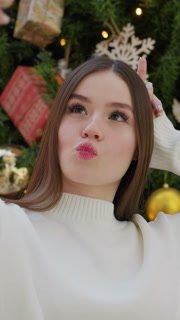 Stylish woman in white top adjusting her phone while taking a playful selfie in front of beautifully decorated Christmas tree with festive