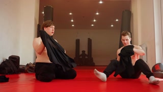 Two men sitting on red mat in gym after training session, wearing their shirts, resting and preparing to change clothes, muscular build