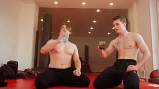 Shirtless young man sitting on red mat inside gym drinking water bottle while another drink from a cup, both appearing tired after intense