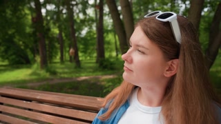 elegant woman on park bench fanning face during sunny day, eyes narrowed, gazing into distance, summer heat relief gesture, white sunglasses