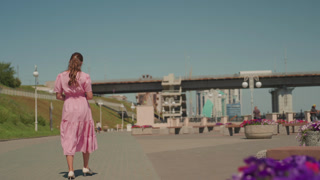 caucasian woman in pink dress walking along sunny urban promenade by bridge and waterfront, pausing near vibrant flower planters while