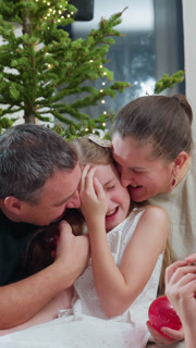 parents kiss daughter near Christmas tree. playful moment shows mother and father whispering, tickling, and gifting ornaments while child