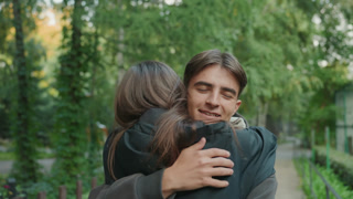 young caucasian couple embrace in park under green trees, backpacks and jackets visible, soft afternoon light, eyes closed in comforting hug