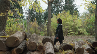 black woman walking past logs in autumn forest, side profile on leafstrewn trail beside stacked timber and tall trees textured bark, muted