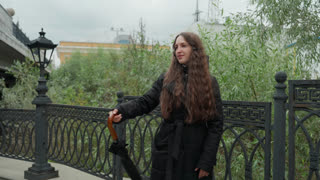 Young lady walking on bridge in city. Smiling woman with curly hair crossing urban riverbank bridge. Cheerful young woman in black coat