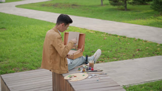 young artist painting sketch in park, sits on wooden bench with portable easel and open paint box palette and brushes scattered nearby