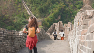 Backpack traveler walking Great Wall stairs, slow paced exploration along ancient stone ramparts, verdant hills in background, distant